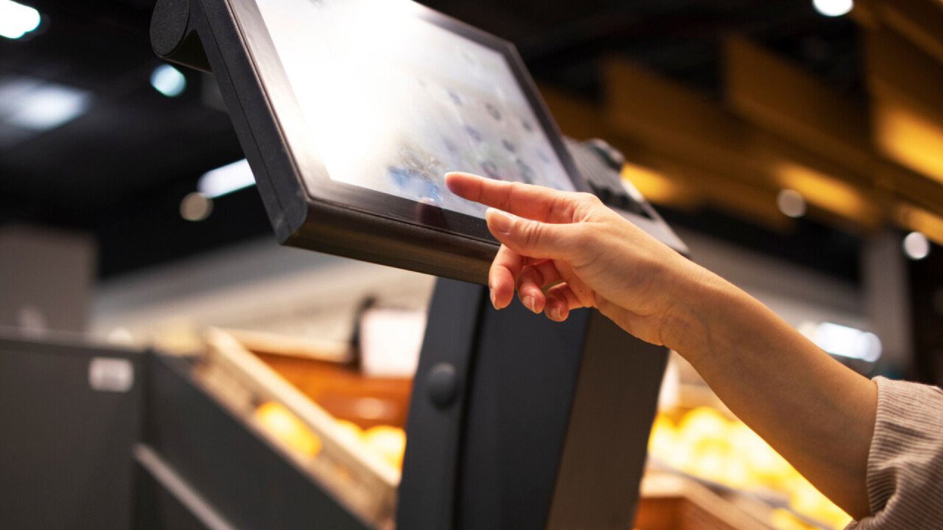 Die Hand einer Person berührt einen Touchscreen-Kiosk in einem Geschäft, wahrscheinlich an einer Selbstbedienungs- oder Kassenstation. Im Hintergrund sind Regale mit unscharfen Artikeln zu sehen.